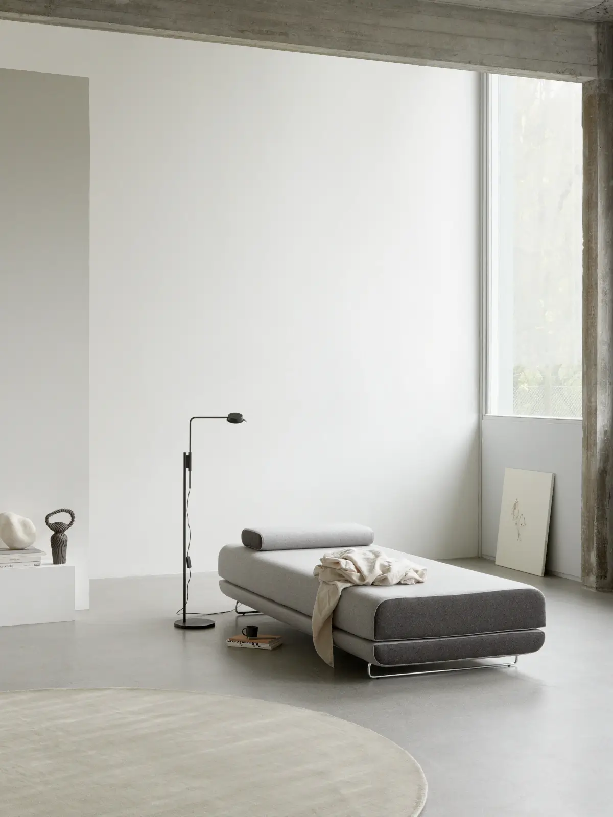 Softline Shine Daybed in Hellgrau mit länglichem Kissen und Metallgestell in einem minimalistischen Wohnambiente mit Stehleuchte, Teppich und großer Fensterfront.