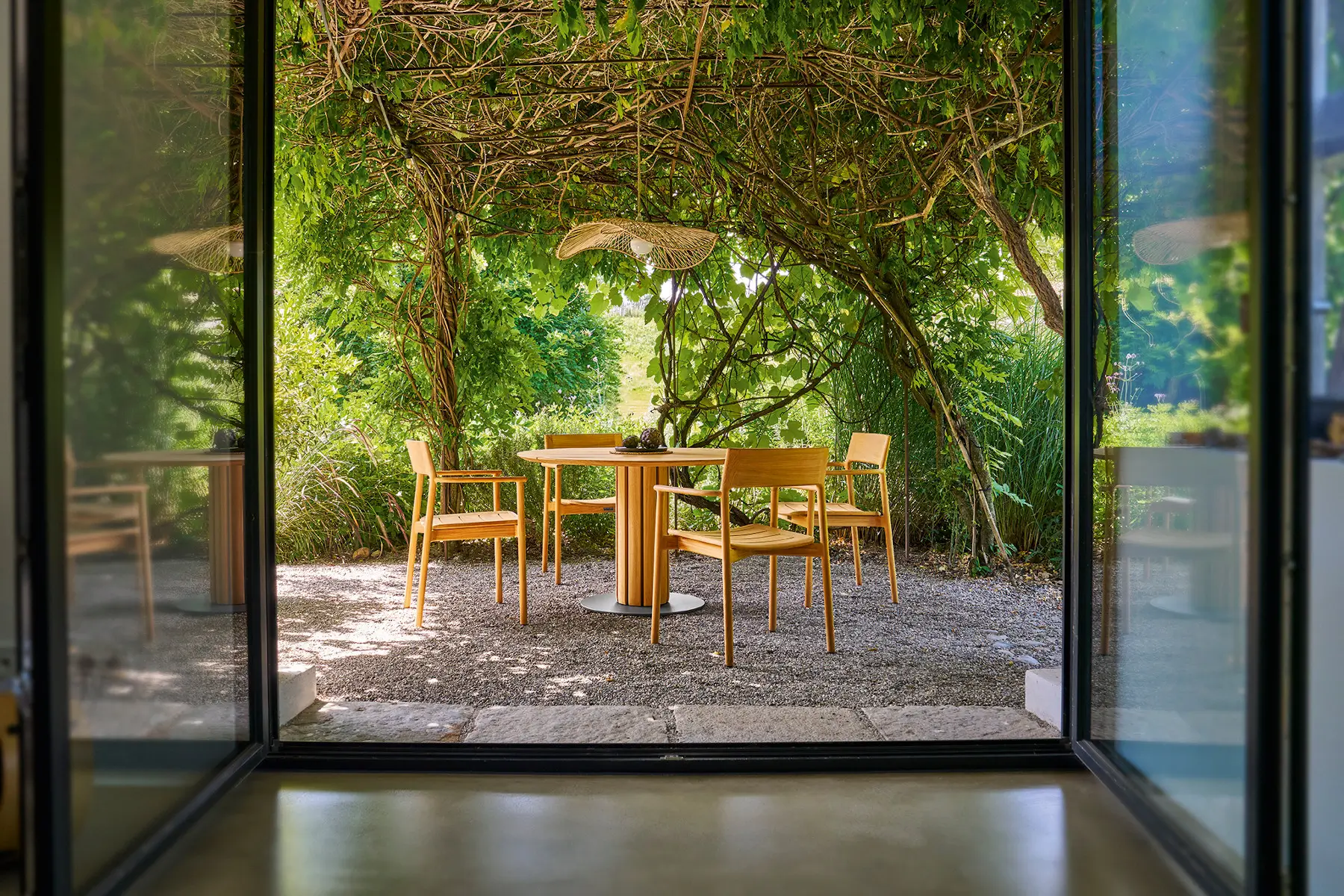 Weishäupl Teva Essgruppe mit rundem Teakholztisch Ø 120 cm und passenden Sesseln in einem grünen Garten. Hochwertige Designmöbel für stilvolle Outdoor-Bereiche.