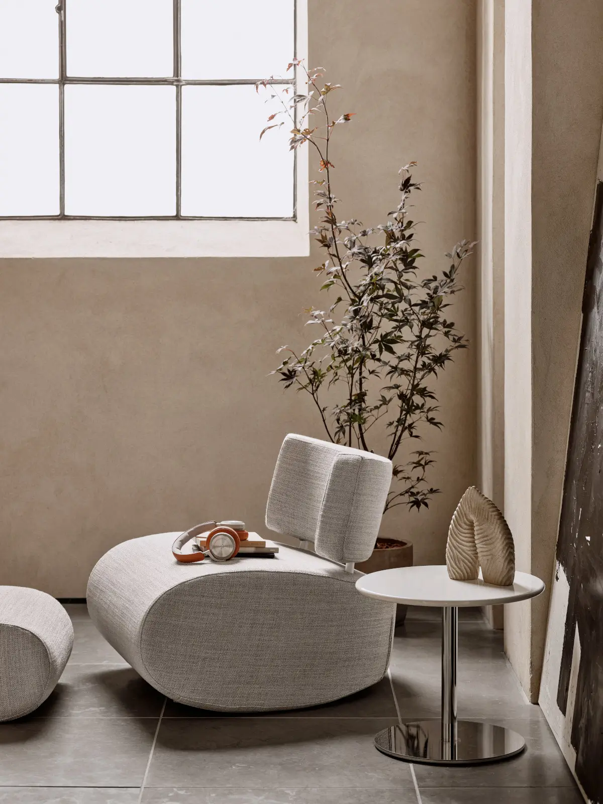 Softline Apollo Sessel in Hellgrau in einem modernen Wohnbereich am Fenster mit passendem Hocker, Pflanze und rundem Beistelltisch.