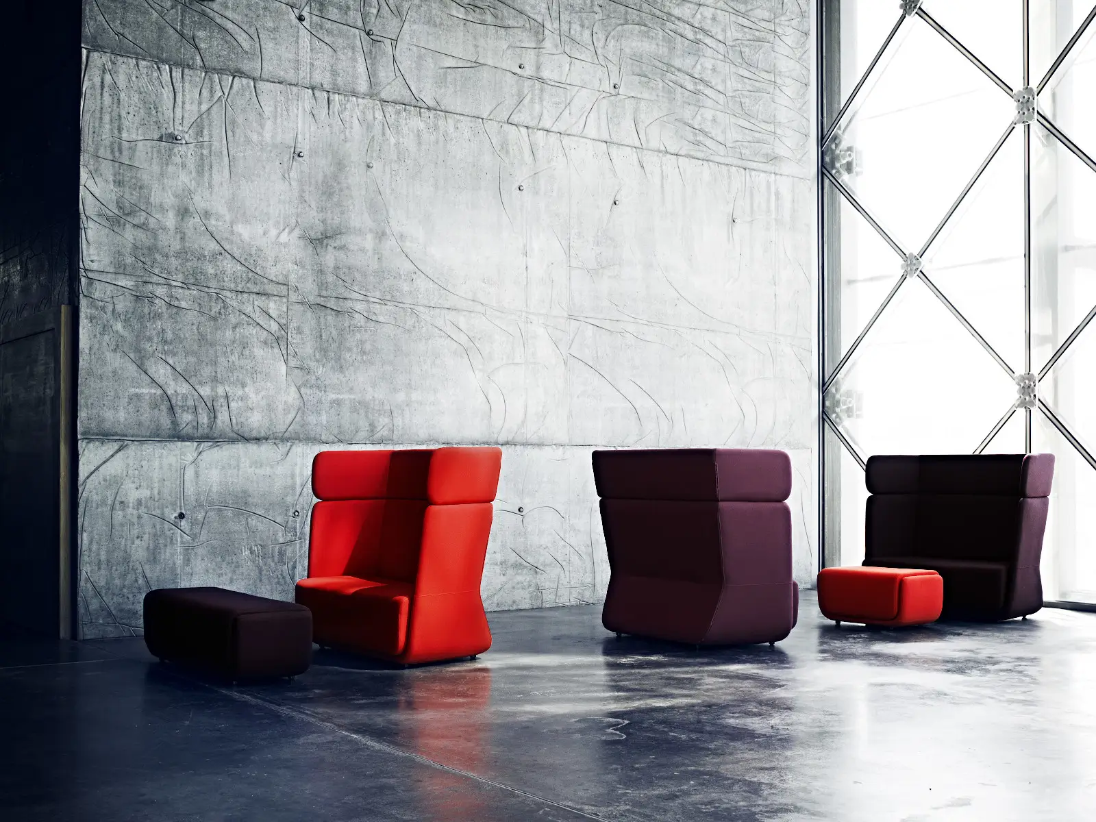 Softline Basket Hocker Small in modernem Lounge-Ambiente mit hohen gepolsterten Sesseln in Rot und dunklen Farbtönen vor Betonwand.