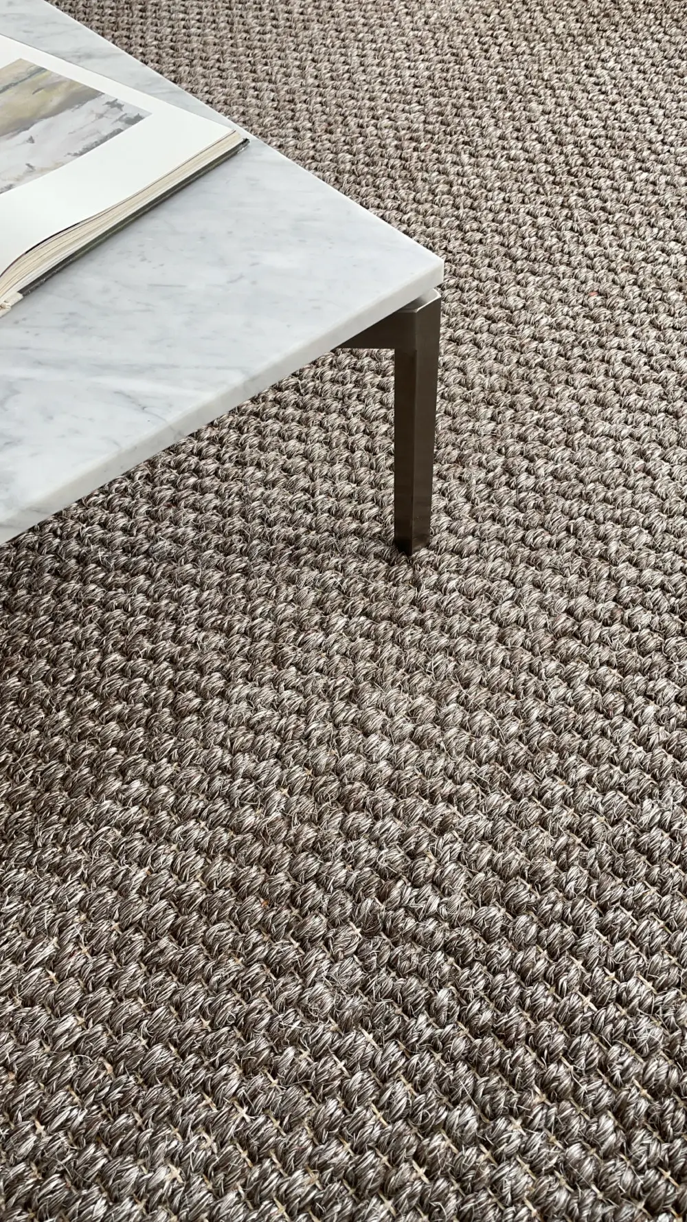 Nahaufnahme des Massimo Copenhagen Belize Teppichs in Taupe mit markanter Webstruktur neben einem Couchtisch mit heller Marmorplatte und Metallgestell.