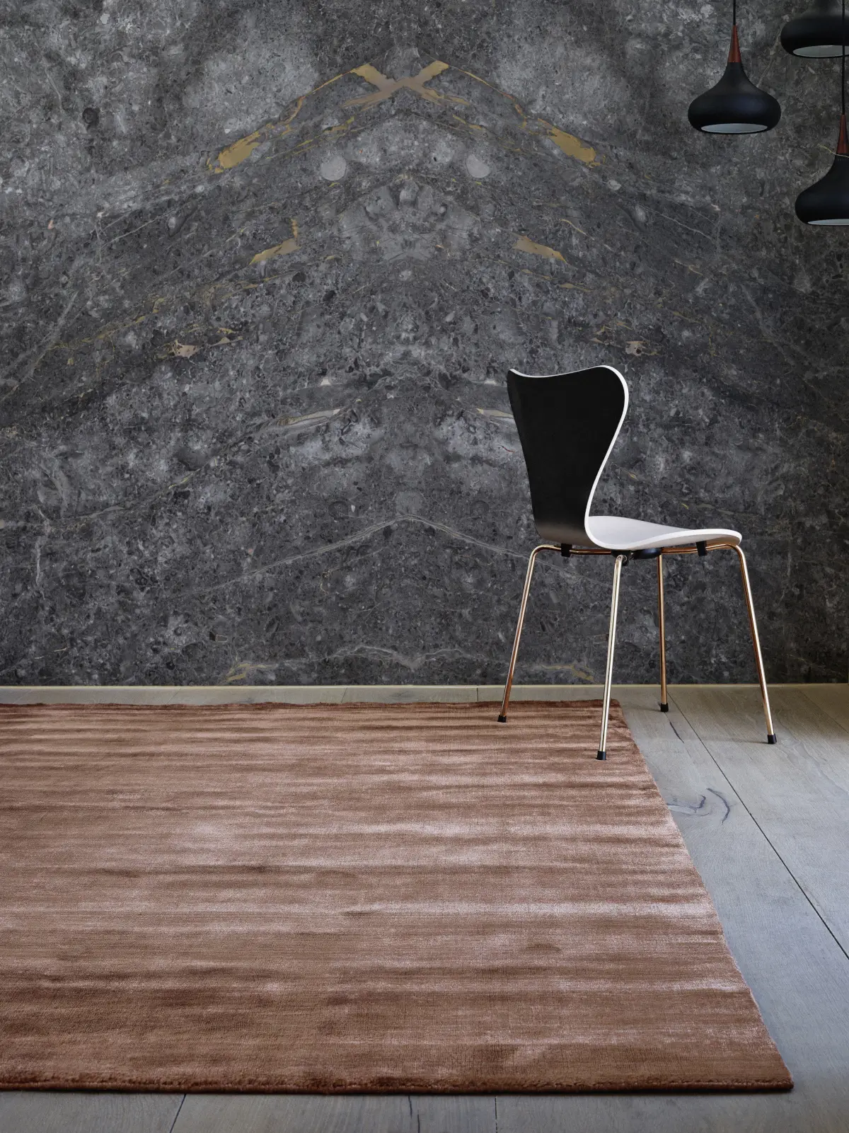 Kupferfarbener Massimo Copenhagen Bamboo Teppich auf hellem Holzboden vor einer dunklen Steinwand mit schwarz-weißem Stuhl und Pendelleuchten.