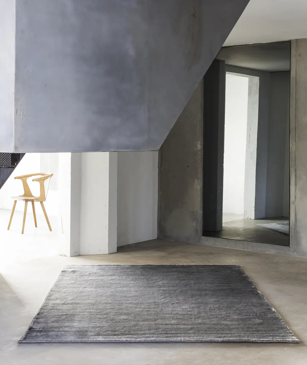 Massimo Copenhagen Bamboo Teppich in Grau auf hellem Betonboden in einem modernen Loft mit Sichtbeton, großem Wandspiegel und Holzstuhl.