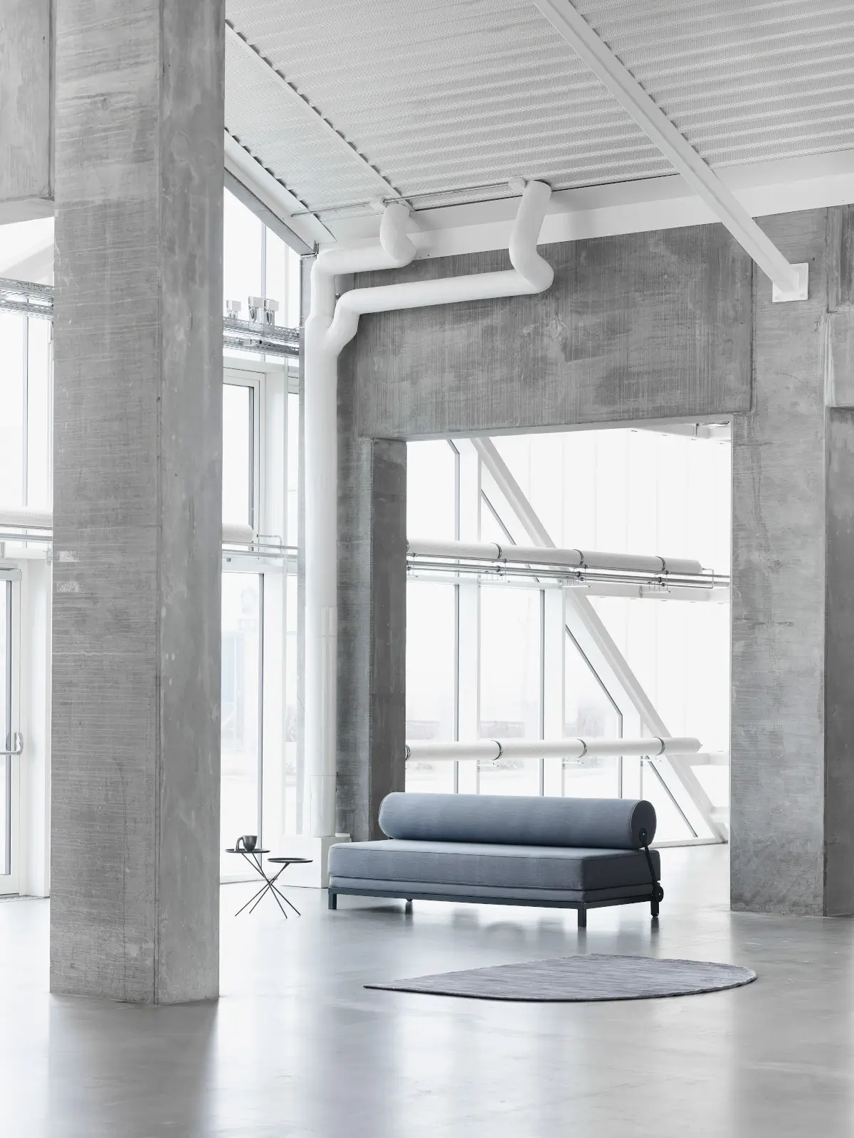 Softline Sleep Sofa in Grau in einem großzügigen modernen Loft mit Betonarchitektur, großer Fensterfront und zylindrischer Rückenlehne.