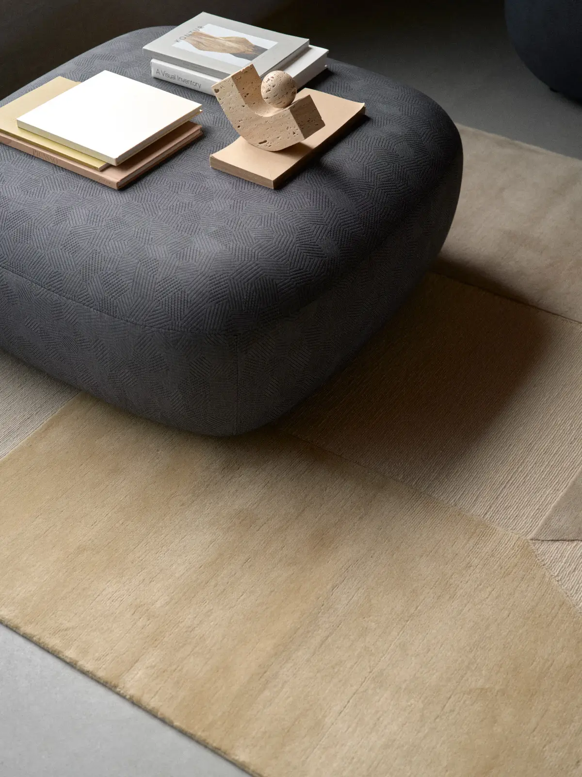 Softline Clay Hocker in Anthrazit aus erhöhter Perspektive mit weich gerundeter Form und strukturierter Stoffoberfläche, dekoriert mit Büchern und Objekt.