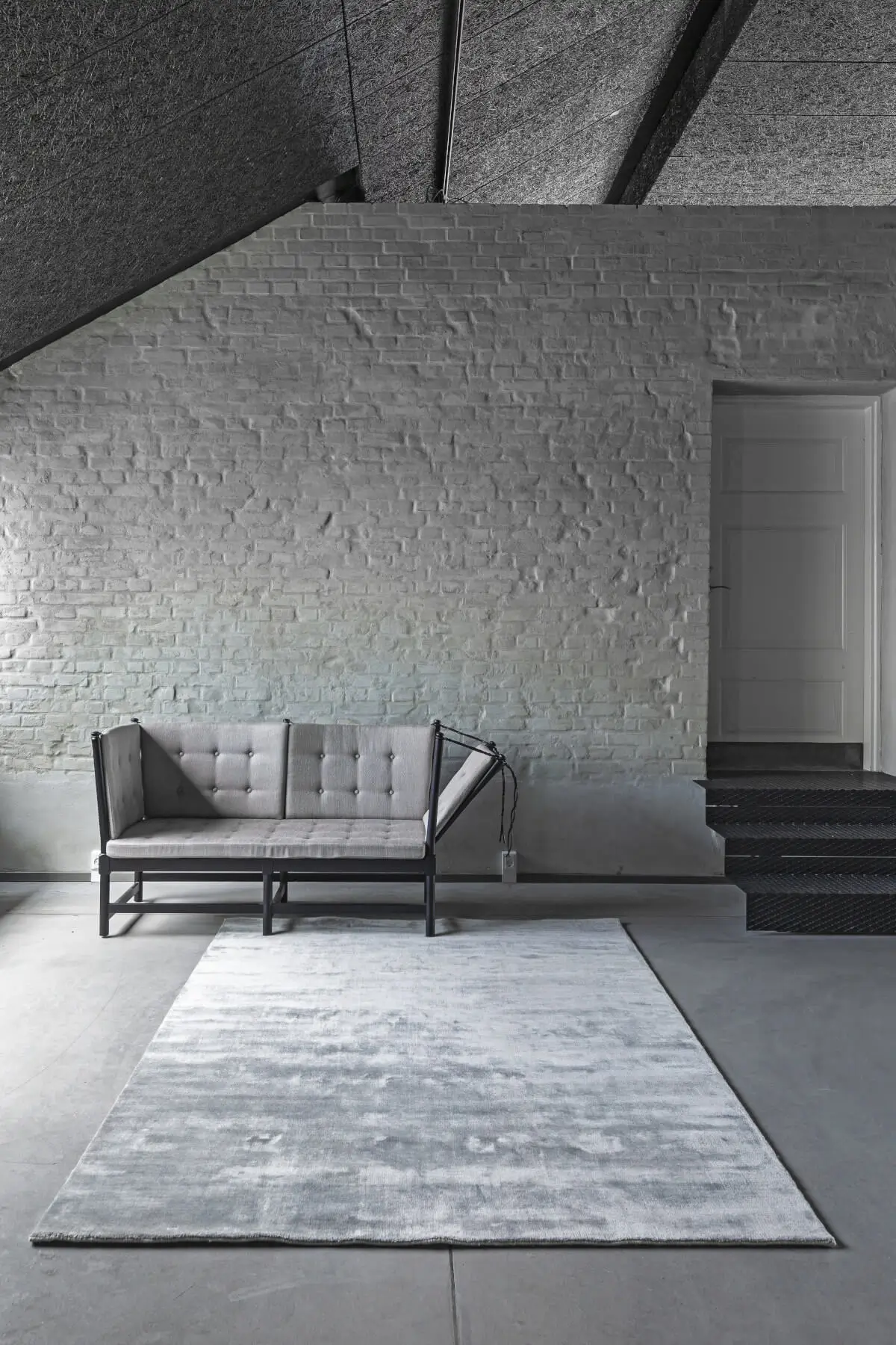 Rechteckiger Teppich in meliertem Grau mit heller und dunkler Schattierung auf einem Betonboden vor einem grauen Sofa und einer weißen Backsteinwand.