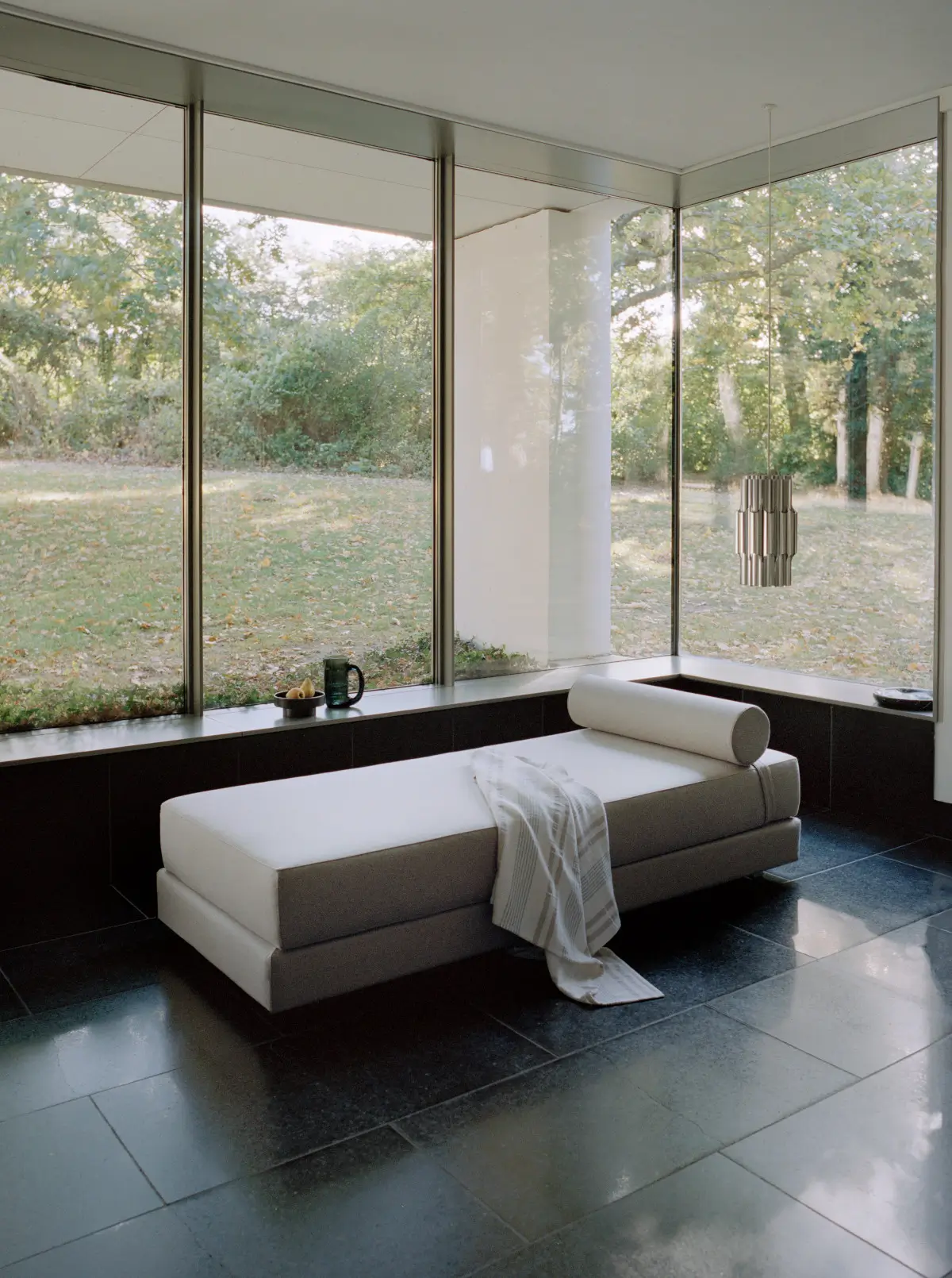 Softline Lubi Daybed in Beige mit rundem Nackenpolster und Decke in einem modernen Wohnraum mit bodentiefen Fenstern und Blick ins Grüne.