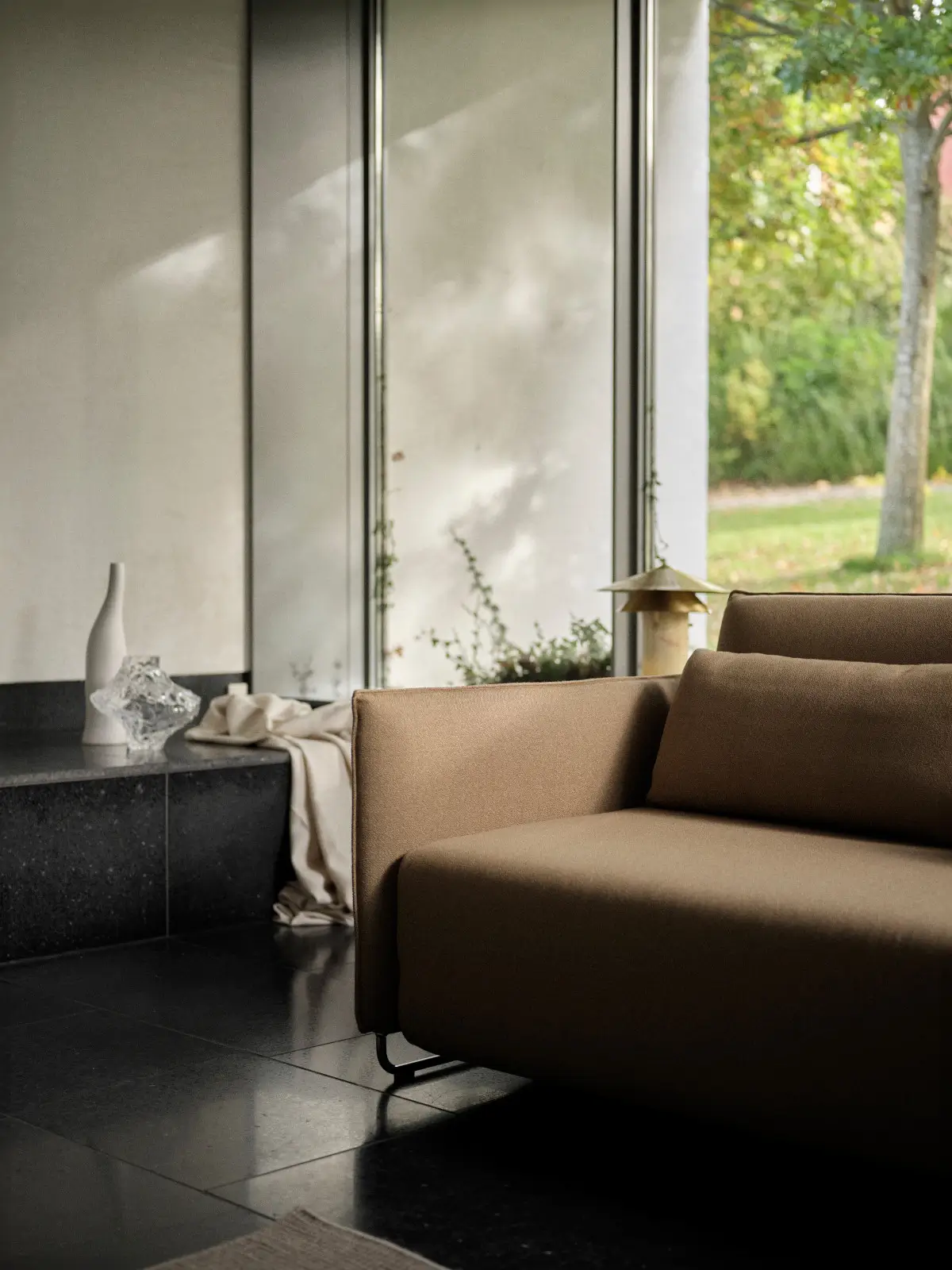 Softline Cord Sofa in Braun mit Rückenkissen und schlanken Metallfüßen in einem modernen Wohnambiente mit großen Fenstern und dunklem Fliesenboden.