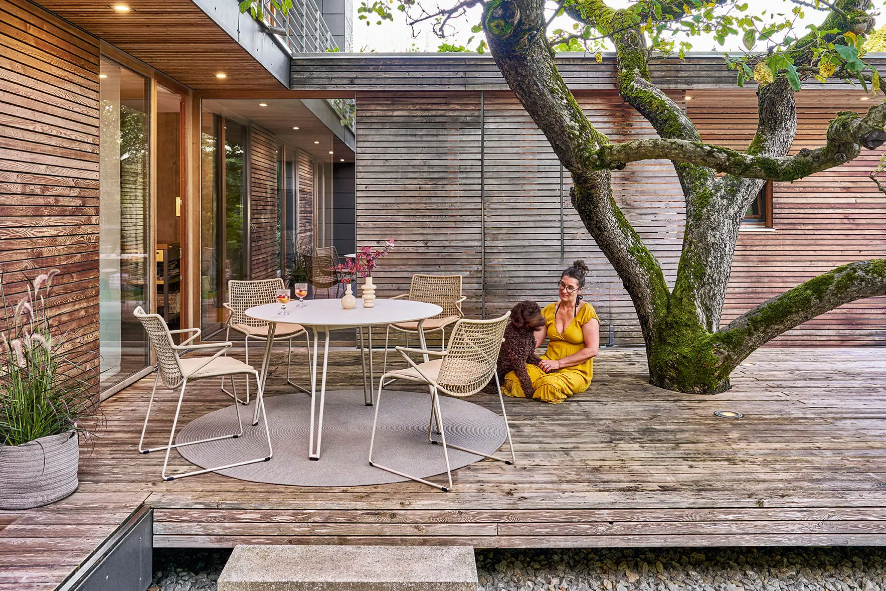 Weishäupl Slope Sitzgruppe in Perlbeige mit rundem Esstisch und Gartenstühlen auf einer modernen Holzterrasse. Hochwertige Outdoor-Designmöbel für Garten und Terrasse.