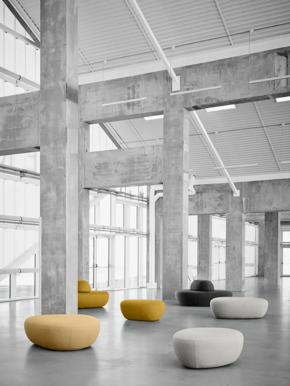 Mehrere Softline Sisters Hocker in Gelb, Grau und Hellgrau in verschiedenen Größen in einem weitläufigen Industrieambiente mit Betonarchitektur.