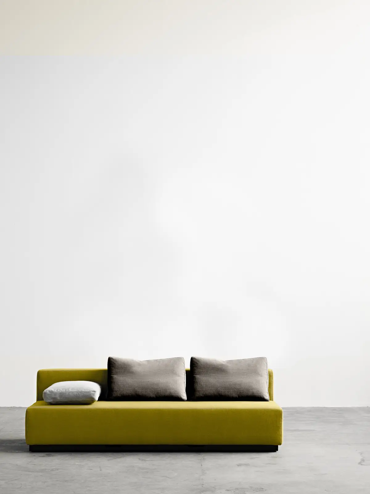 Softline Nevada Sofa 3-Sitzer in Gelb aus frontaler Perspektive mit zwei dunkelgrauen Rückenkissen, hellem Seitenkissen und schwarzem Sockel.