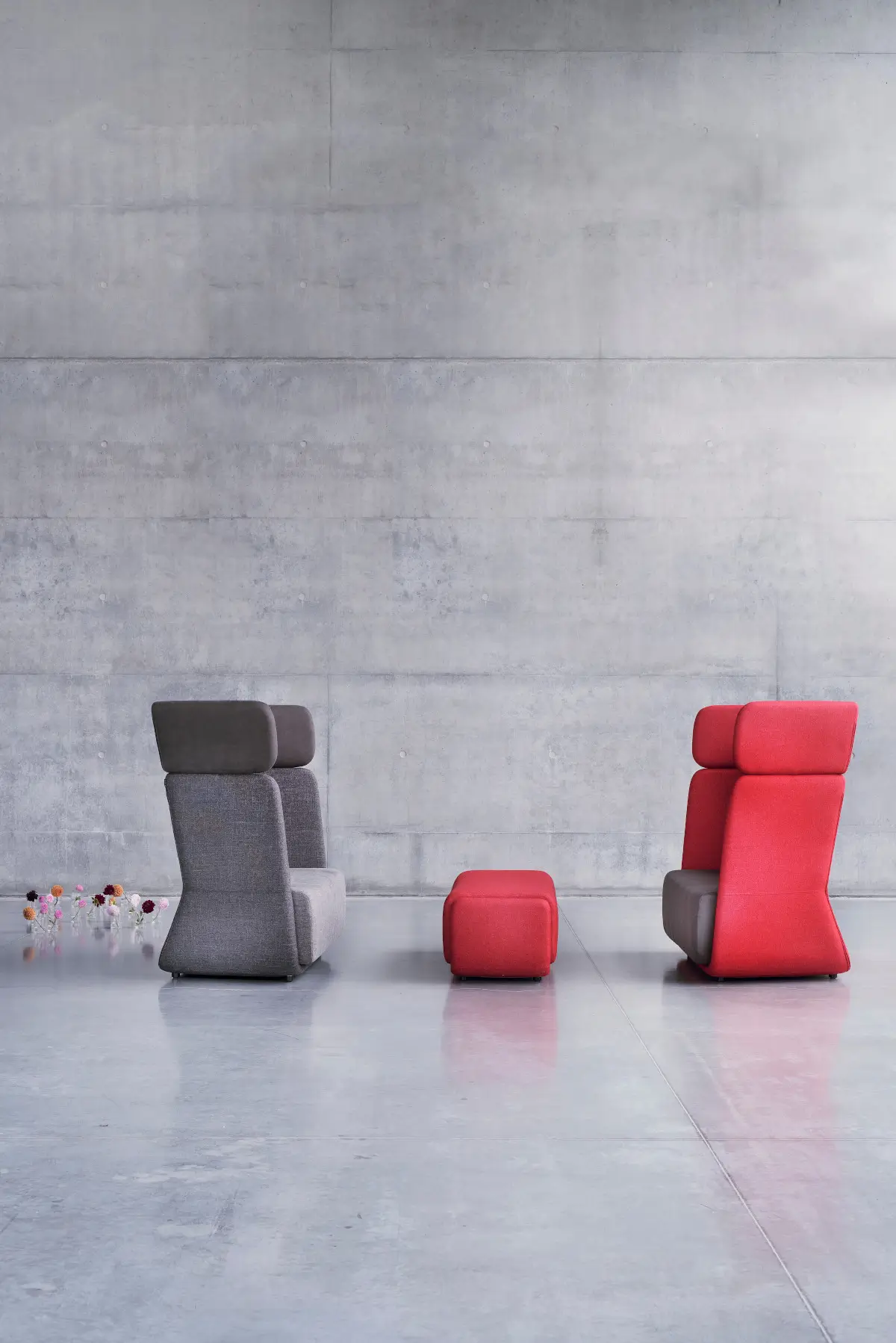 Softline Basket Hocker Small in Rot zwischen zwei hohen gepolsterten Sesseln in Grau und Rot vor einer Betonwand.