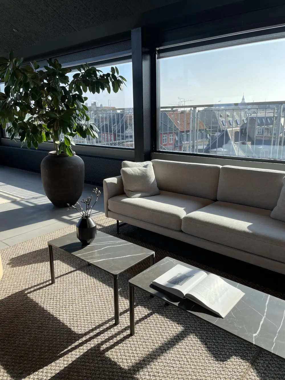 Massimo Copenhagen Belize Teppich in Natural unter zwei schwarzen Couchtischen mit Marmoroptik vor einem grauen Sofa in einem modernen, lichtdurchfluteten Wohnzimmer.