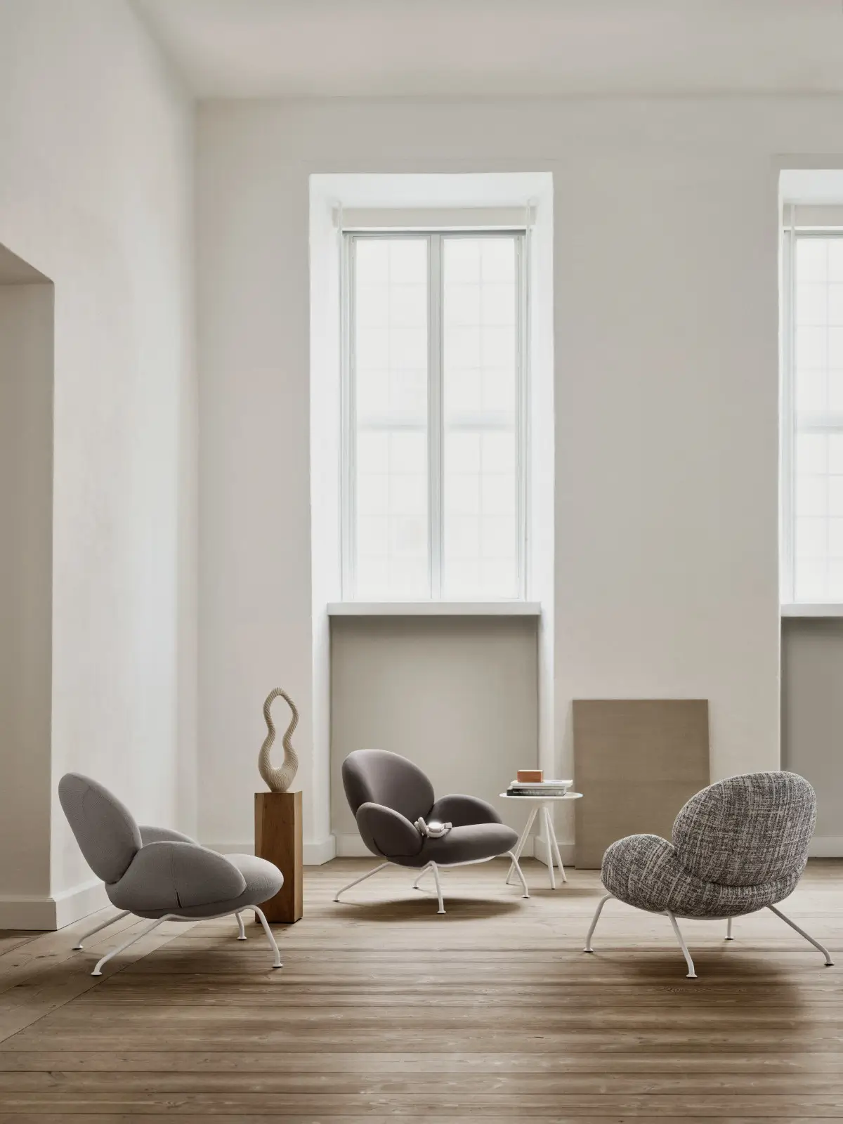 Drei Softline Baixa Sessel in Grau und gemustertem Grau mit weißen Metallbeinen in einem hellen Wohnraum mit Holzboden und großen Fenstern.