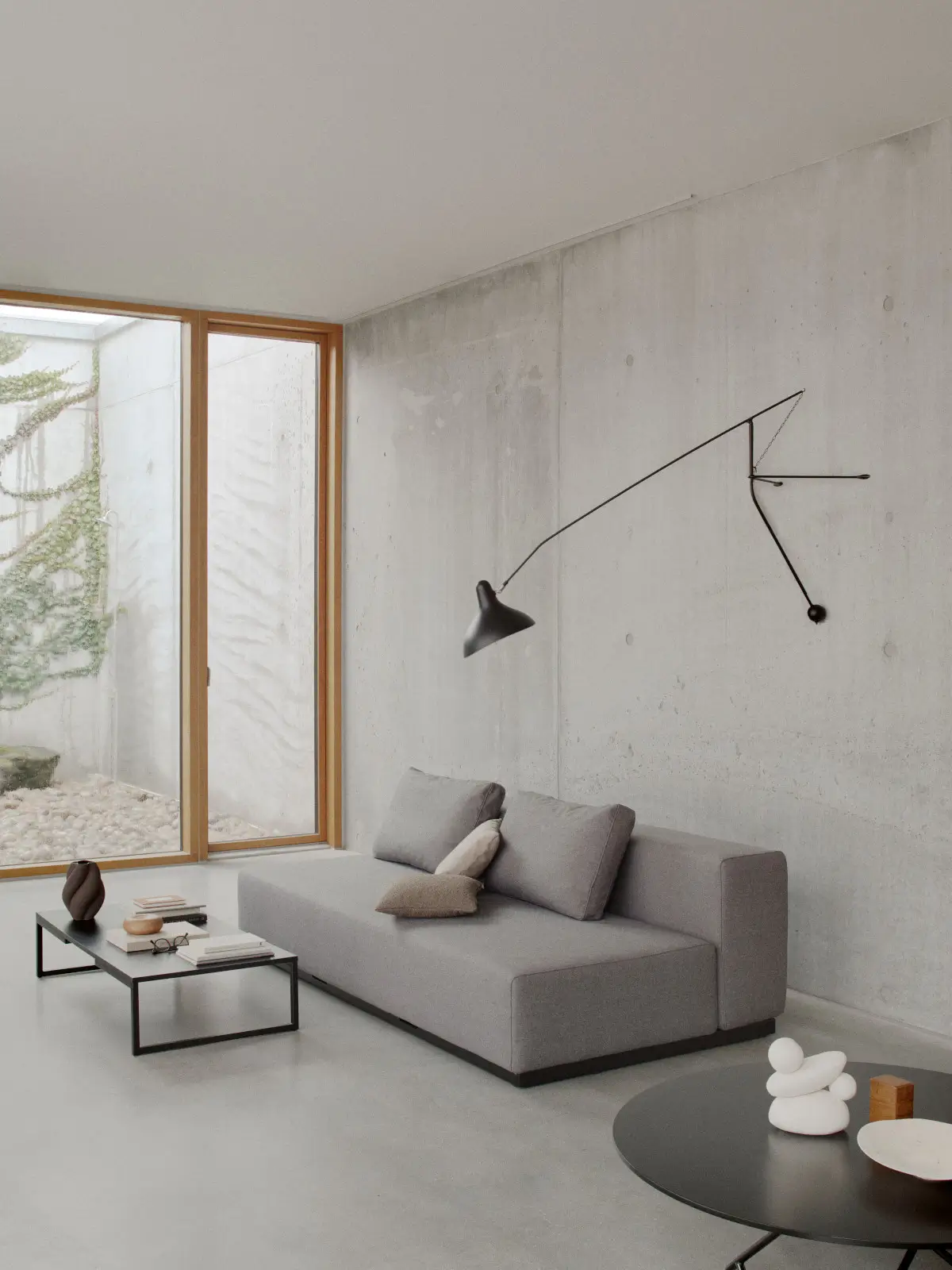 Softline Nevada Sofa 3-Sitzer in Grau mit Rückenkissen und zusätzlichen Dekokissen in einem minimalistischen Wohnambiente mit Betonwand und großen Fenstern.