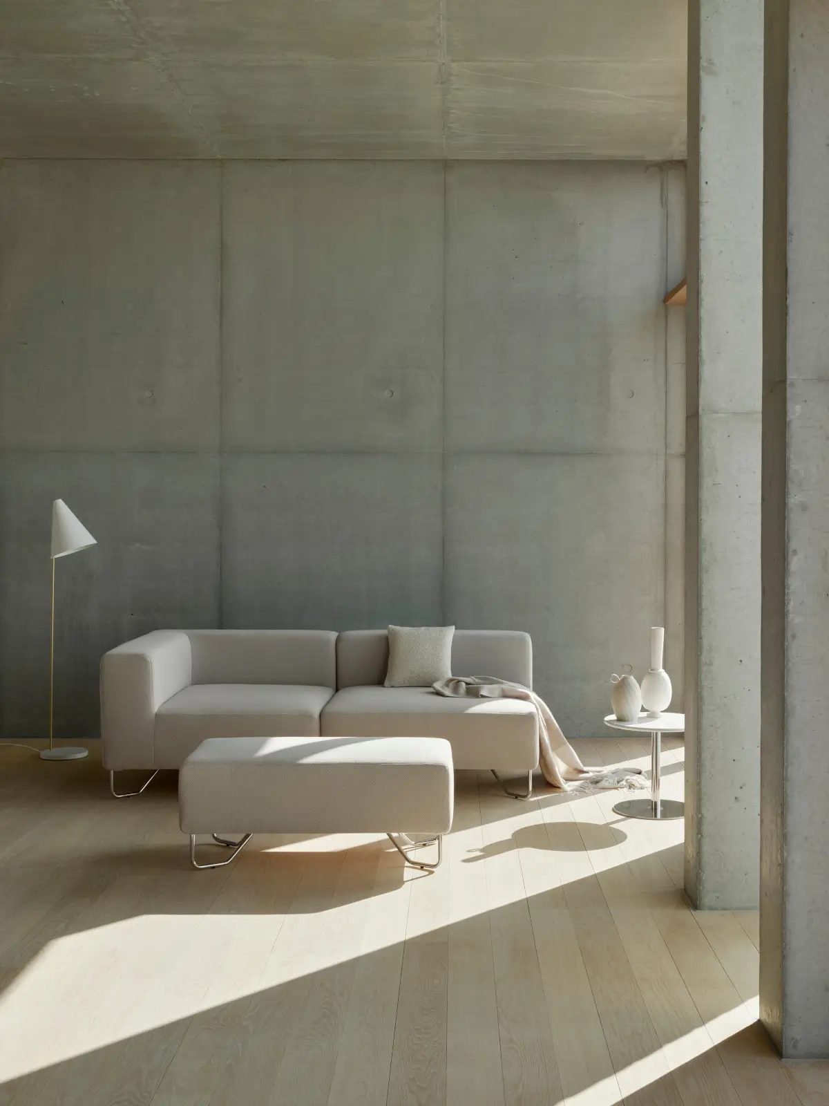 Softline Lotus Sofa in Beige mit passendem Hocker in einem modernen Wohnambiente mit Betonwänden, hellem Holzboden, Stehleuchte und Beistelltisch.