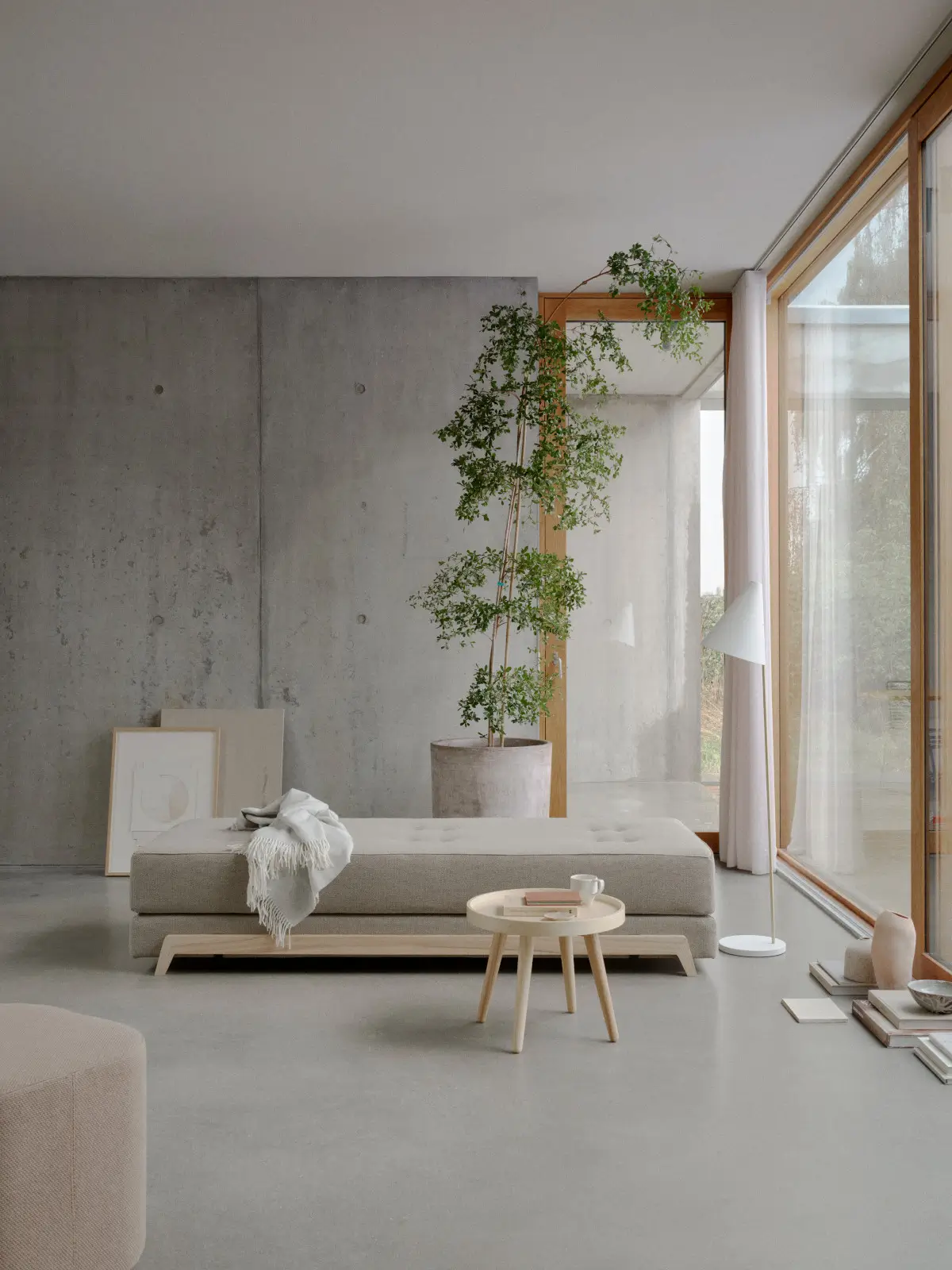 Softline Frame Daybed in Beige in einem modernen Wohnambiente mit großen Fenstern, Betonwand, Pflanze und rundem Holztisch.