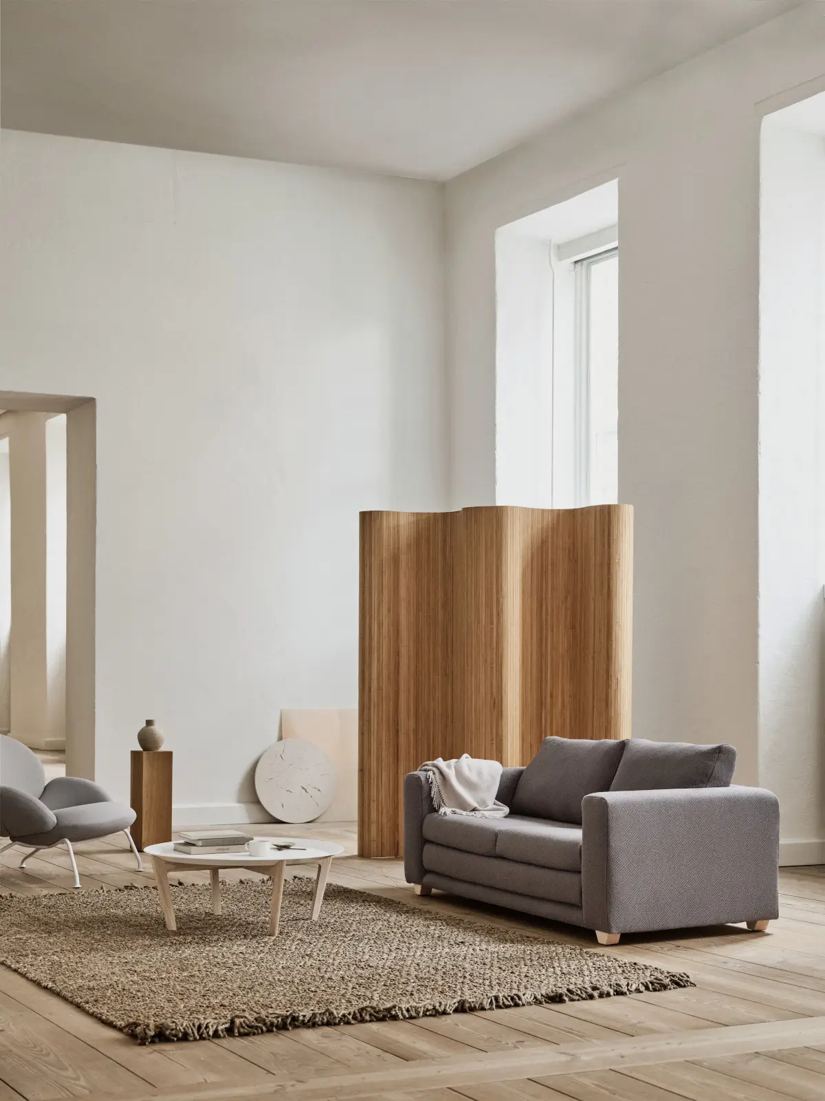 Softline Victor Sofa 2,5-Sitzer in Grau mit Kissen und Decke in einem hellen Wohnambiente mit Naturteppich, Holzboden, Couchtisch und Holz-Raumteiler.