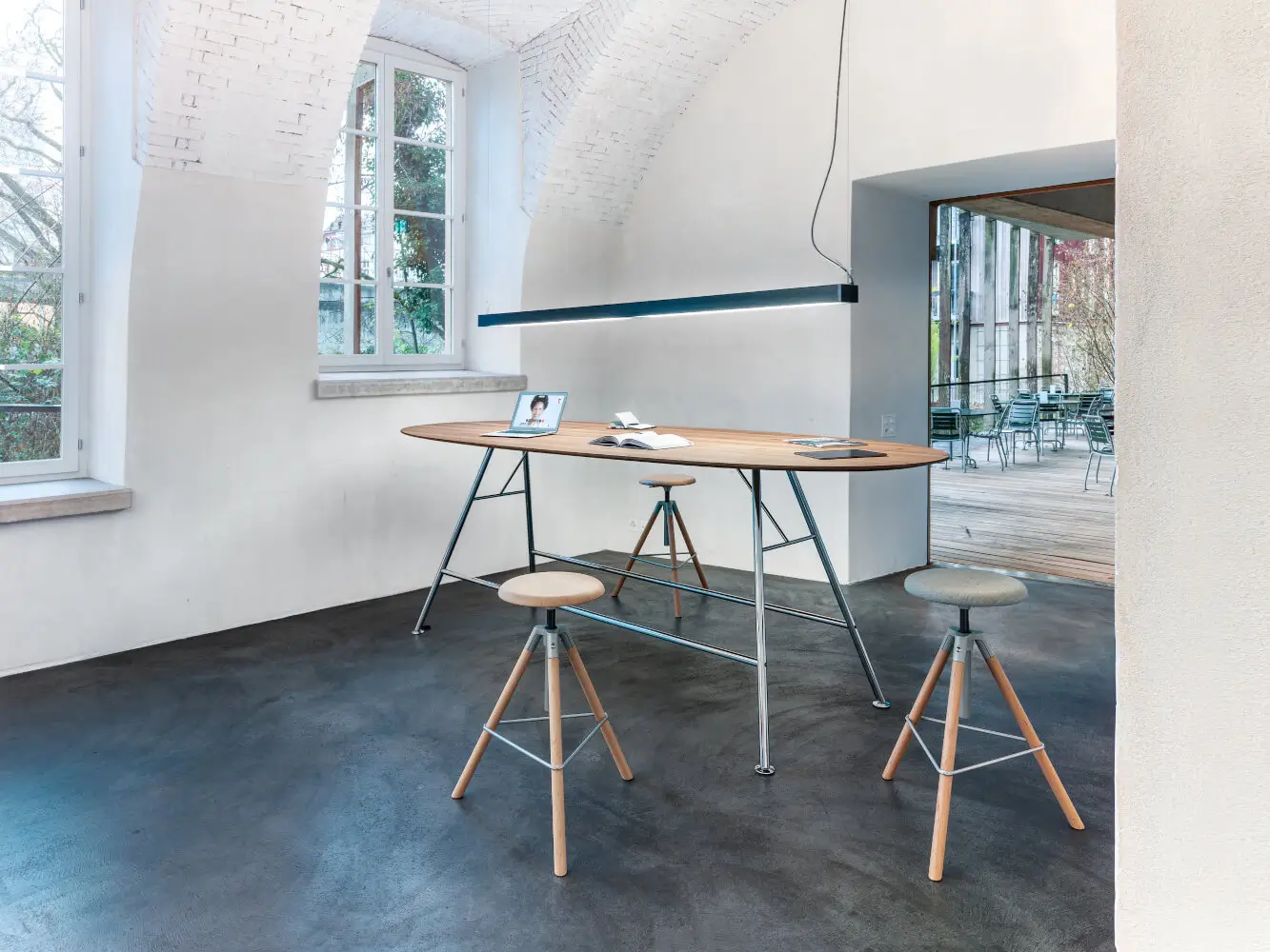 Girsberger Akio Steel Stehtisch mit ovaler Massivholzplatte in einem modernen Arbeitsbereich mit Hockern und Pendelleuchte.