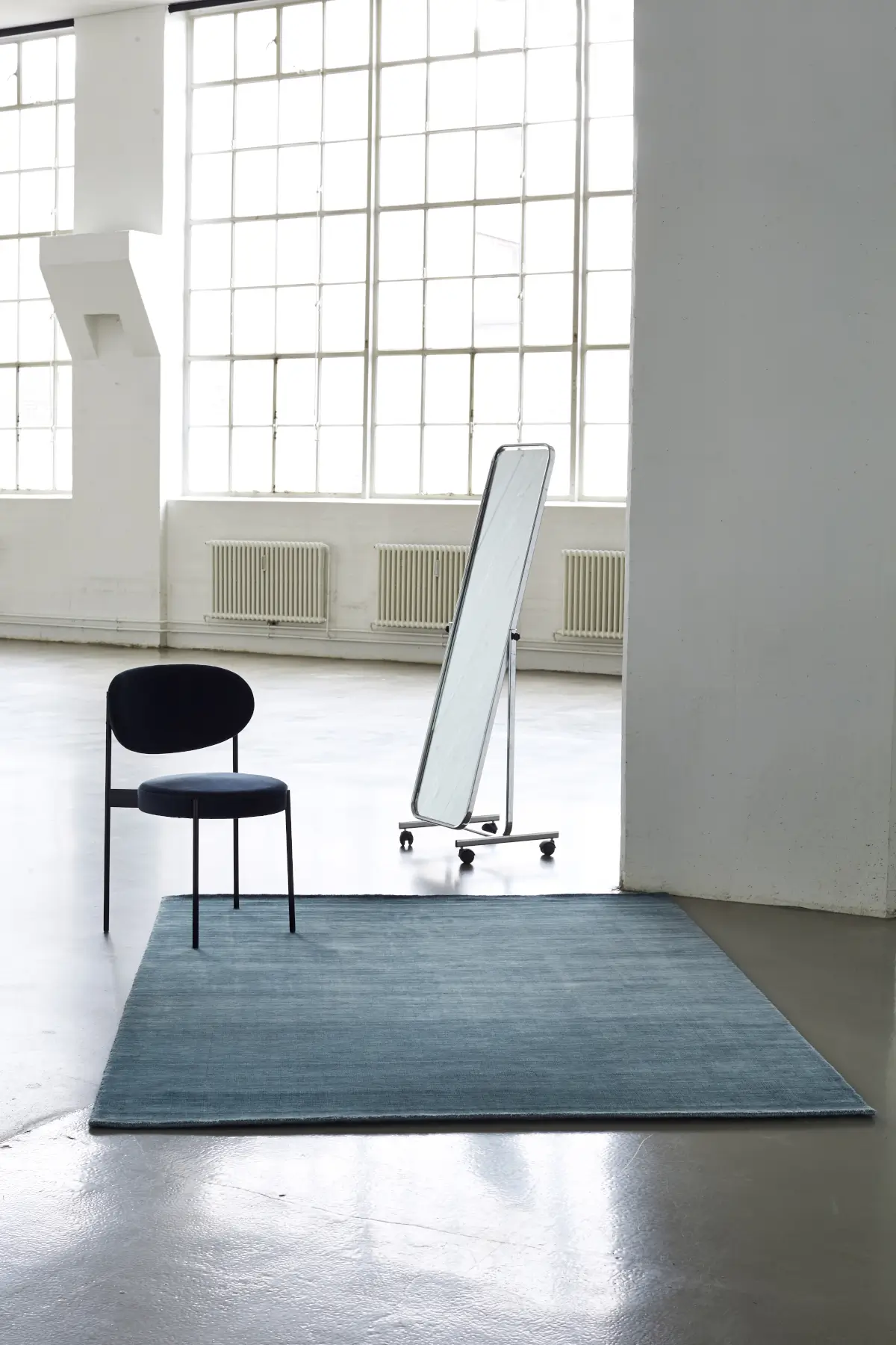 Massimo Copenhagen Bamboo Teppich in Petrol auf hellem Boden in einem lichtdurchfluteten Loft mit schwarzem Stuhl, Standspiegel und großen Fenstern.