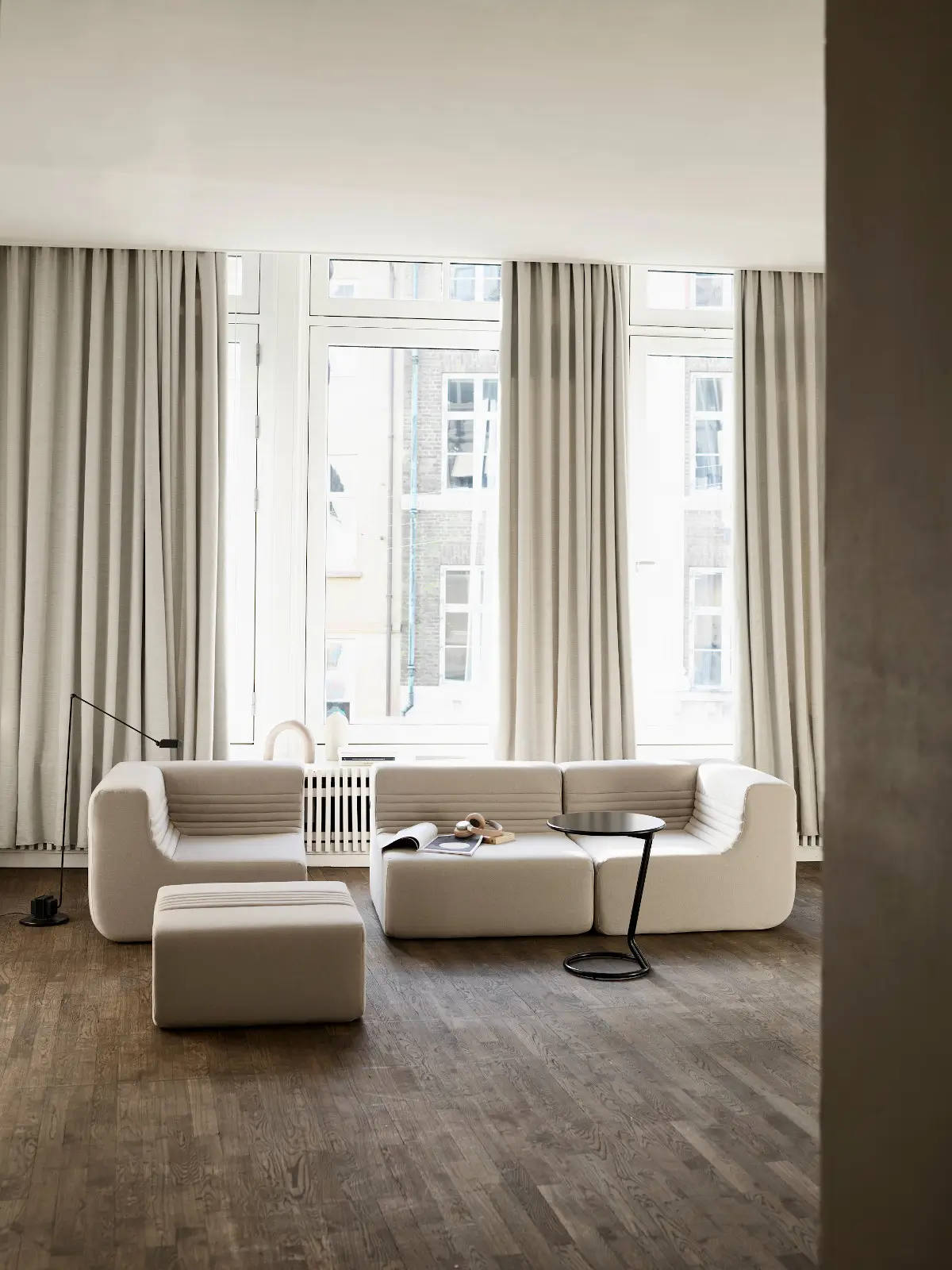 Softline Loft Sofa in Weiß mit mehreren Modulen und passendem Hocker in einem hellen Wohnraum mit großen Fenstern, Holzboden und Beistelltisch.