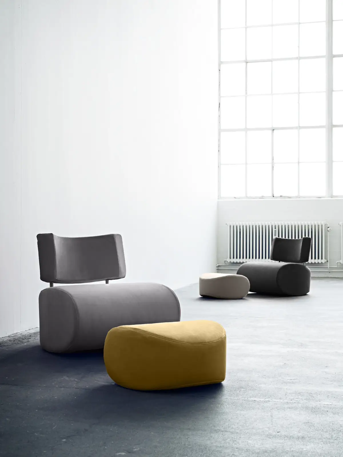 Softline Apollo Hocker in Gelb vor einem grauen Sessel in einem hellen modernen Wohnambiente; im Hintergrund stehen weitere Sitzmöbel und ein Hocker.