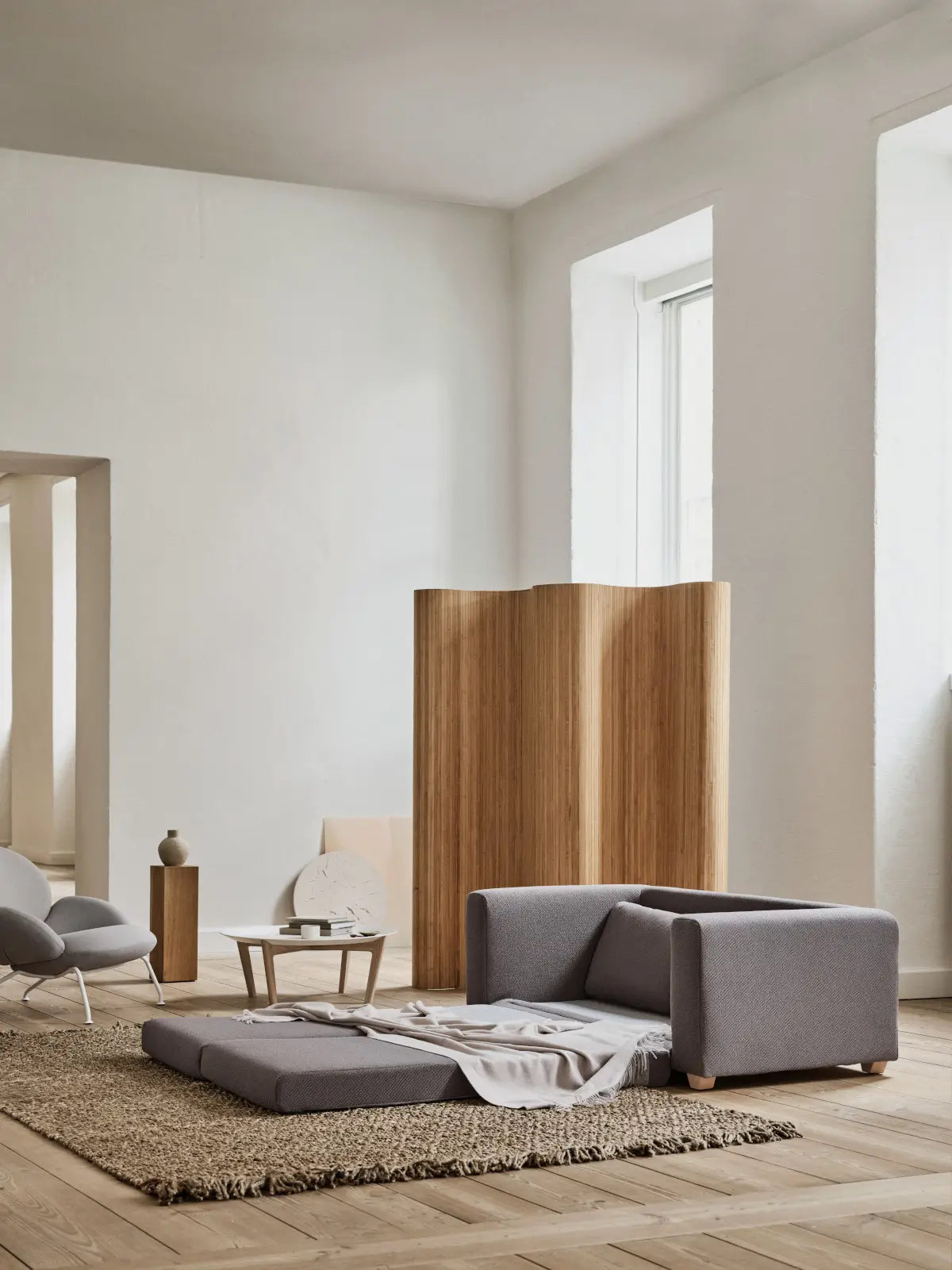 Softline Victor Sofa 2,5-Sitzer in Grau mit ausgezogener Liegefläche in einem hellen Wohnambiente mit Holzboden, Teppich und Raumteiler aus Holz.