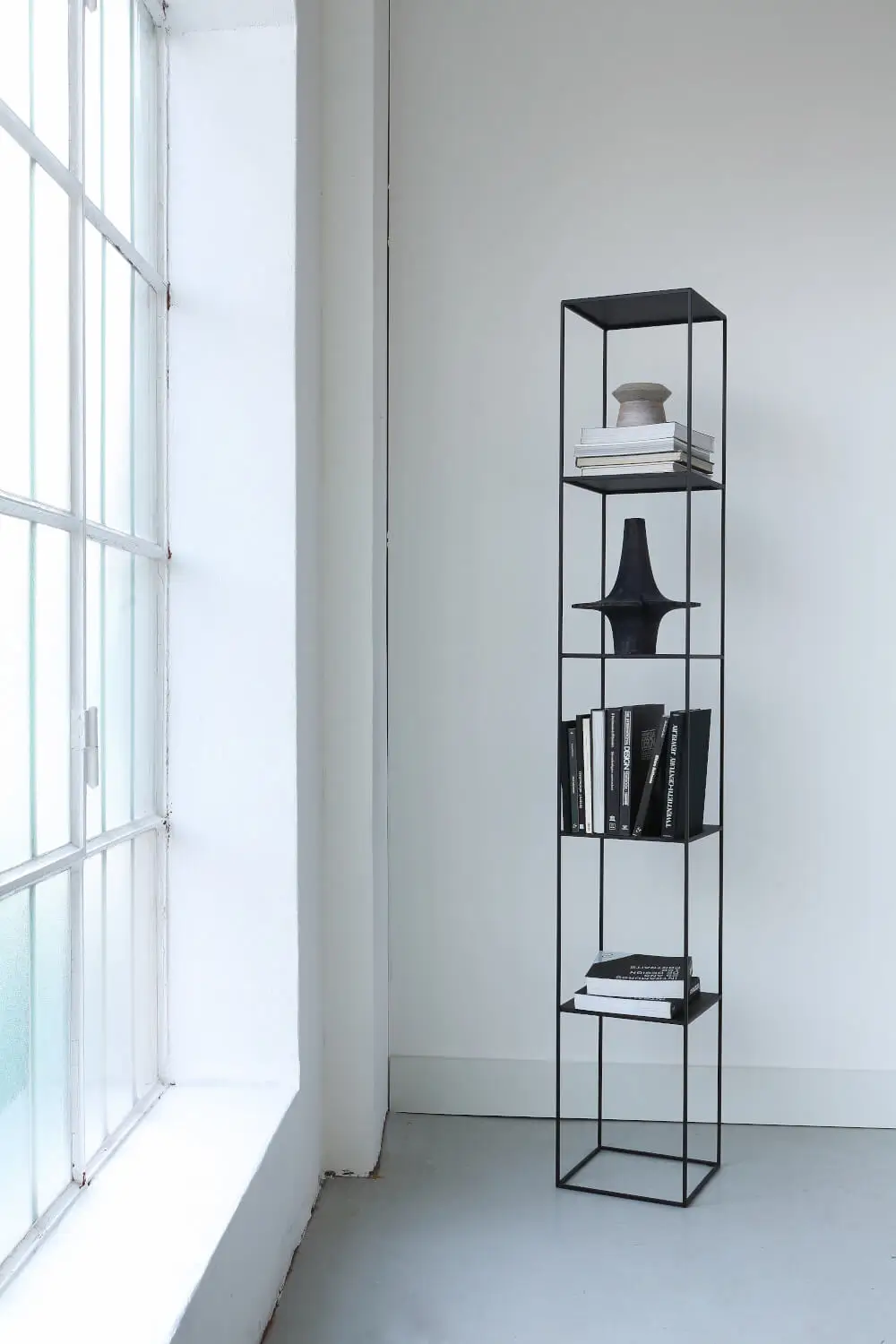Zeus Slim Irony Column Regal mit Büchern und Wohnaccessoires neben einer großen Fensterfront.