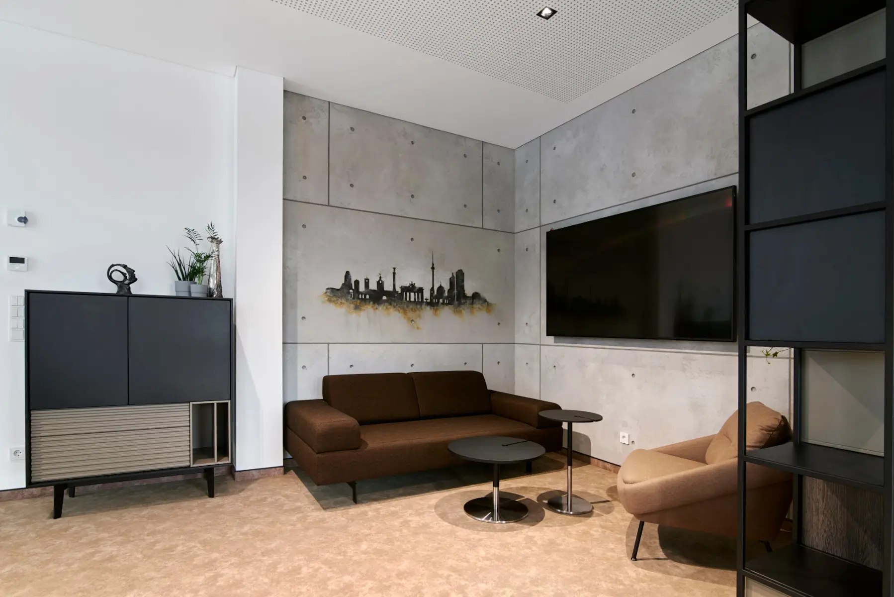 Beiger Noti Nome Sessel mit dunklen Holzbeinen in einem modernen Büro-Sitzbereich mit braunem Sofa, Couchtischen und Betonwänden