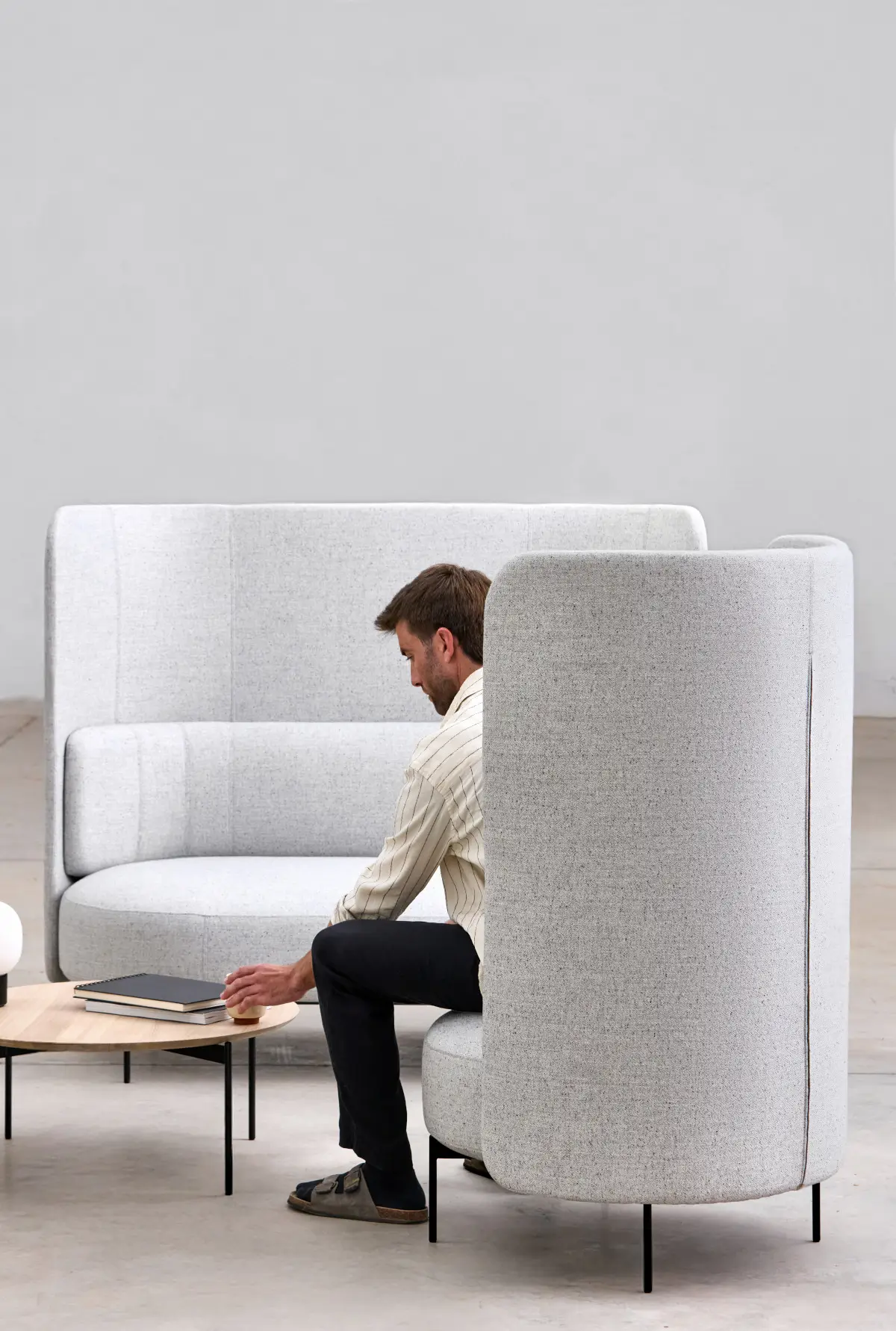 Softline Joe Sofa Hoch in Grau im Lounge-Ambiente mit passendem Hochlehner-Sessel, Couchtisch und einer sitzenden Person.