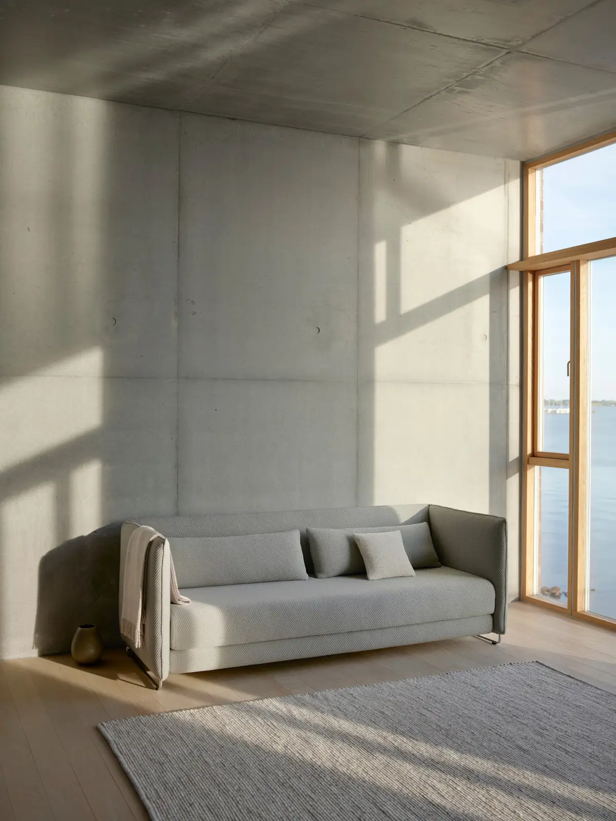 Softline Metro Sofa in Hellgrau in einem modernen Wohnambiente mit großen Fenstern, Betonwänden, hellem Holzboden und Teppich.