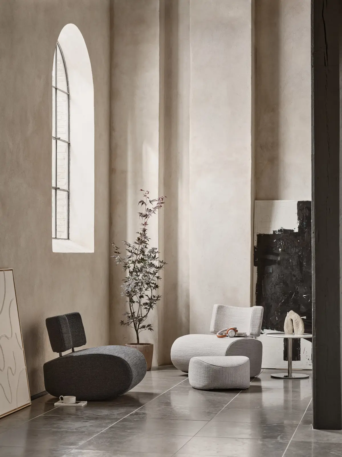Softline Apollo Hocker in Hellgrau vor einem passenden Sessel in einem modernen Wohnbereich mit hohen Fenstern, Pflanzen und Beistelltisch.