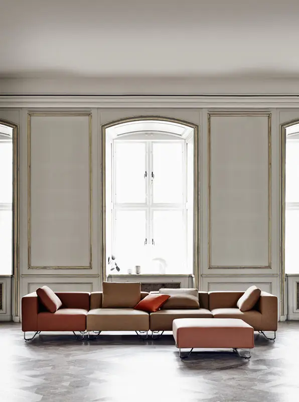 Softline Lotus Hocker in Braun vor einem modularen Sofa in Braun und Beige in einem eleganten, hellen Wohnambiente mit hohen Fenstern.