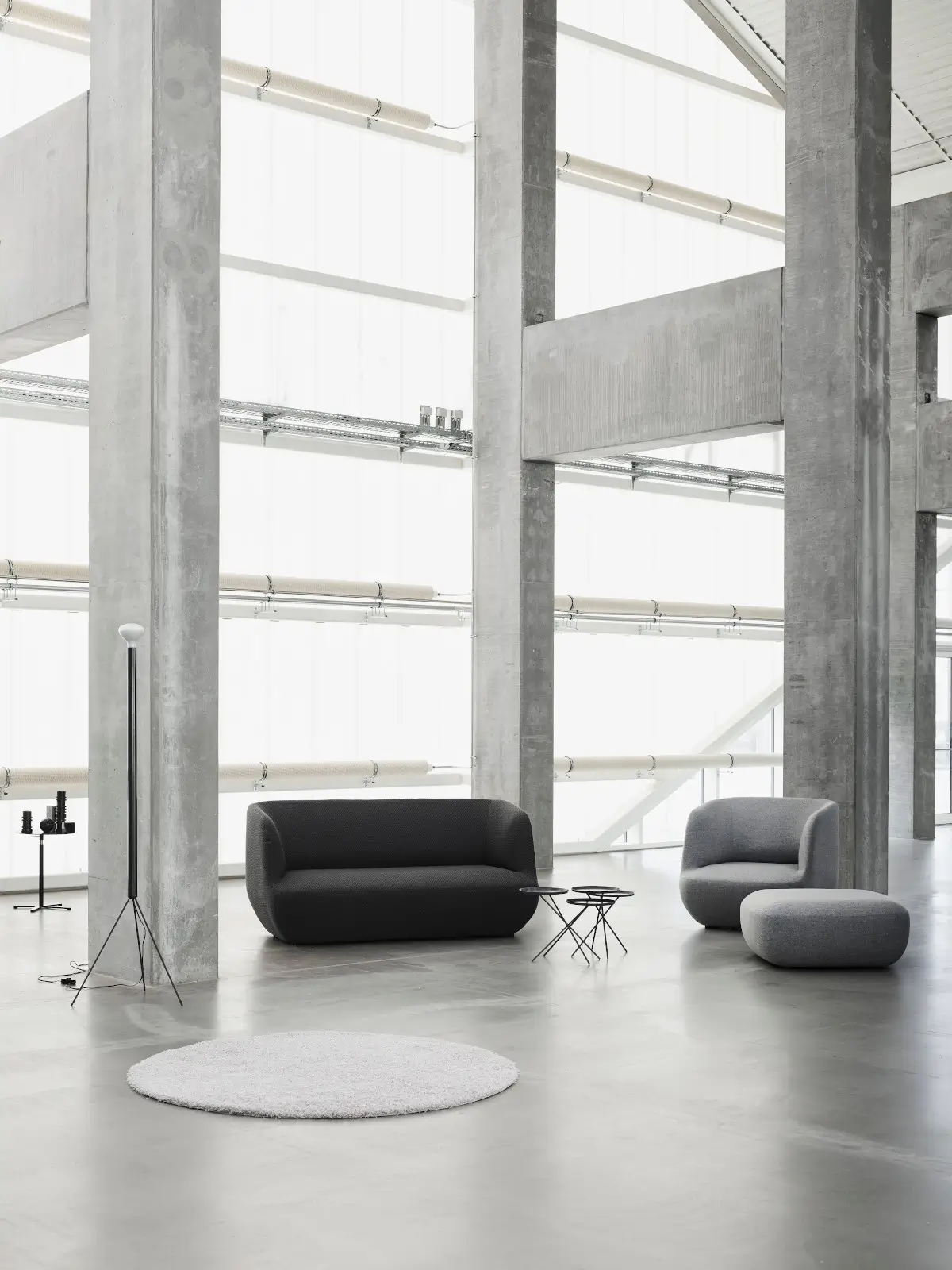 Softline Clay Hocker in Grau neben passendem Sessel und Sofa in einer großzügigen, modernen Lounge mit Betonarchitektur.